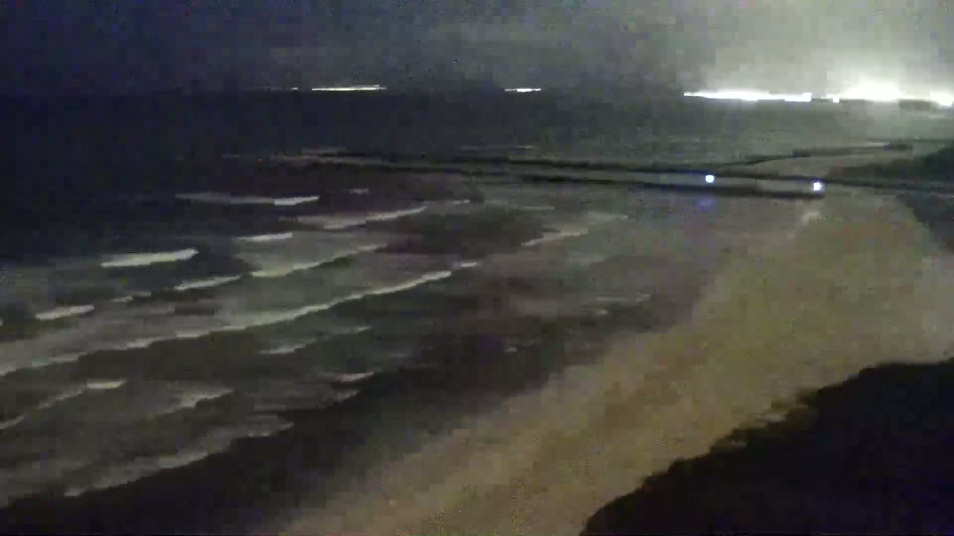 A dark, nighttime view of a beach with waves gently lapping the shore under a cloudy sky.