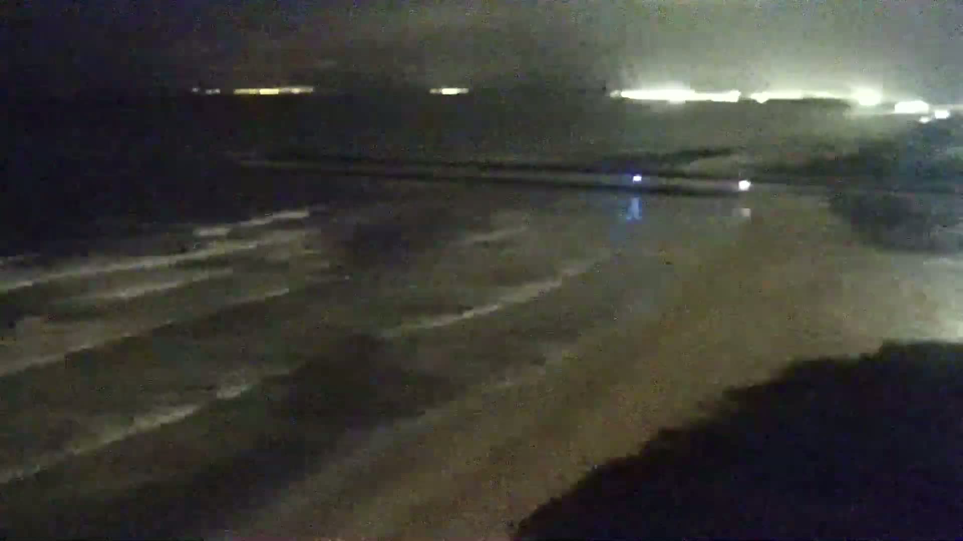 A dark, nighttime beach scene shows gentle waves lapping the shore under a cloudy sky, with distant city lights faintly visible on the horizon.