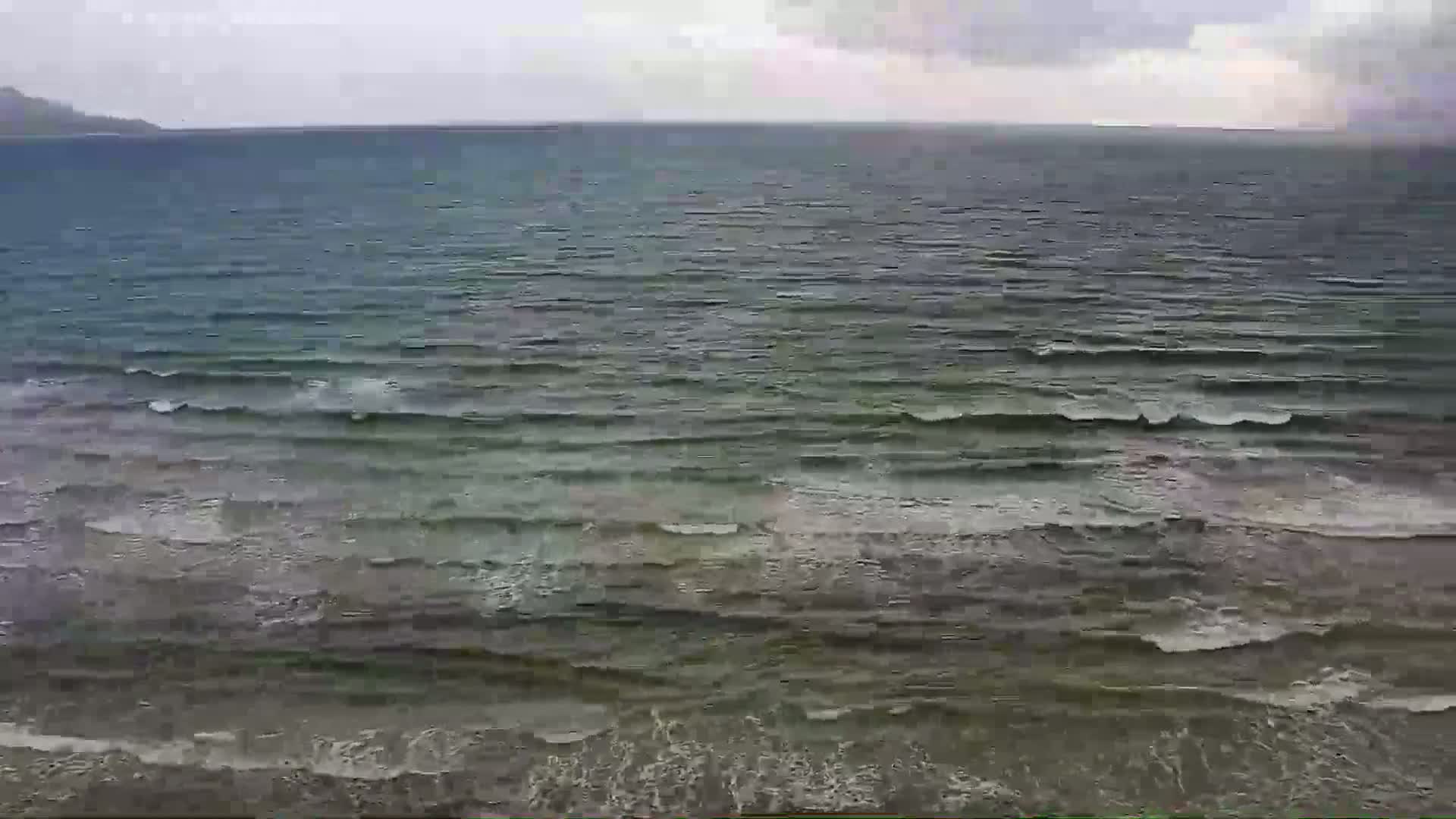 A body of water with small waves under an overcast sky shows a distant shoreline.