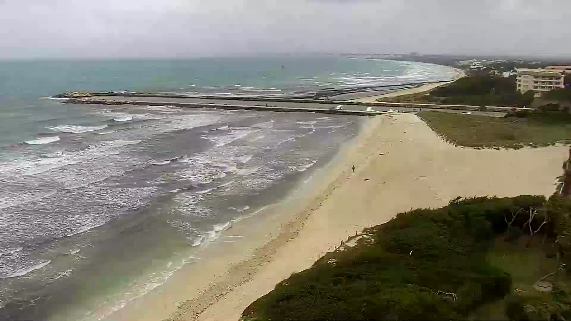 A long sandy beach with moderate waves under an overcast sky is bordered by green vegetation and a distant building.