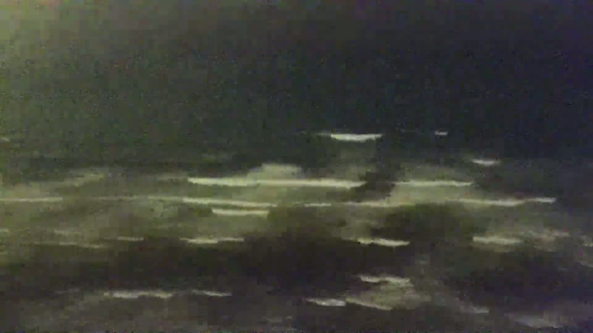 A dark, nighttime scene shows a body of water with faint white streaks suggesting waves or reflections under a mostly dark sky.