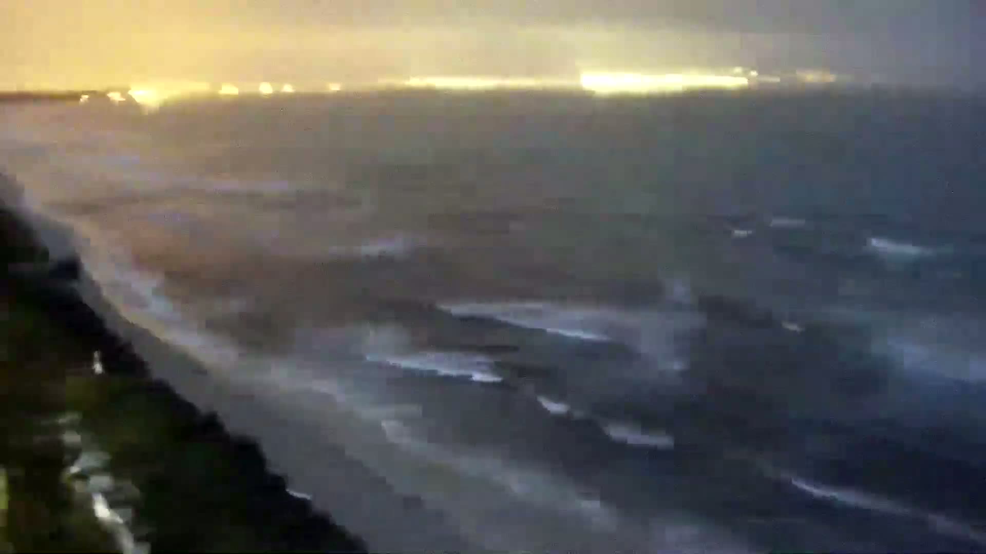 A nighttime coastal scene shows dark, turbulent waves crashing on a shore under a hazy, dimly lit sky.