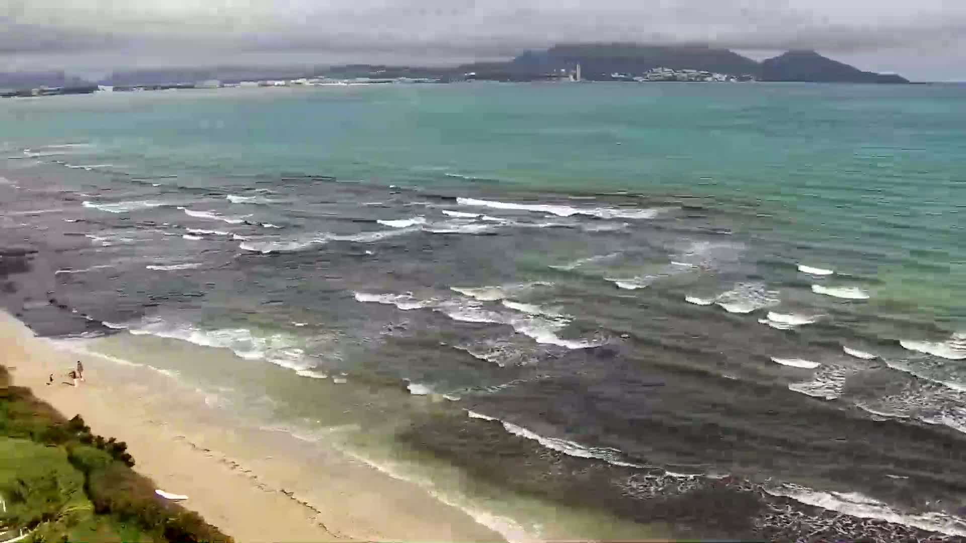 A sandy beach with a few people on it slopes down to a dark-colored ocean with small waves under a cloudy sky, with land and buildings visible in the distance.