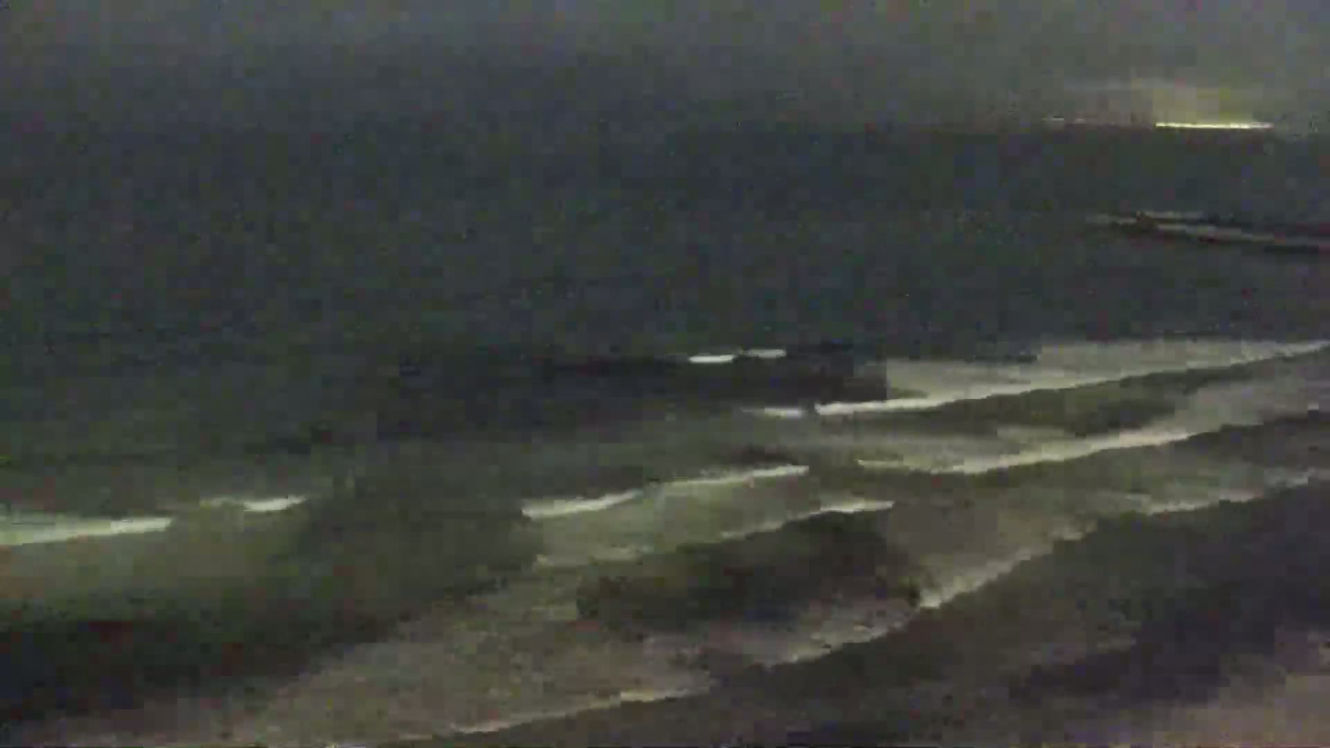Waves crash on a dark, sandy shore under a cloudy, nighttime sky.