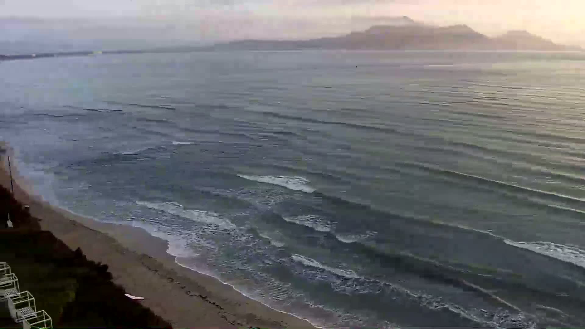 A sandy beach slopes down to a calm, gray sea with small waves under an overcast sky, and distant mountains are visible on the horizon.