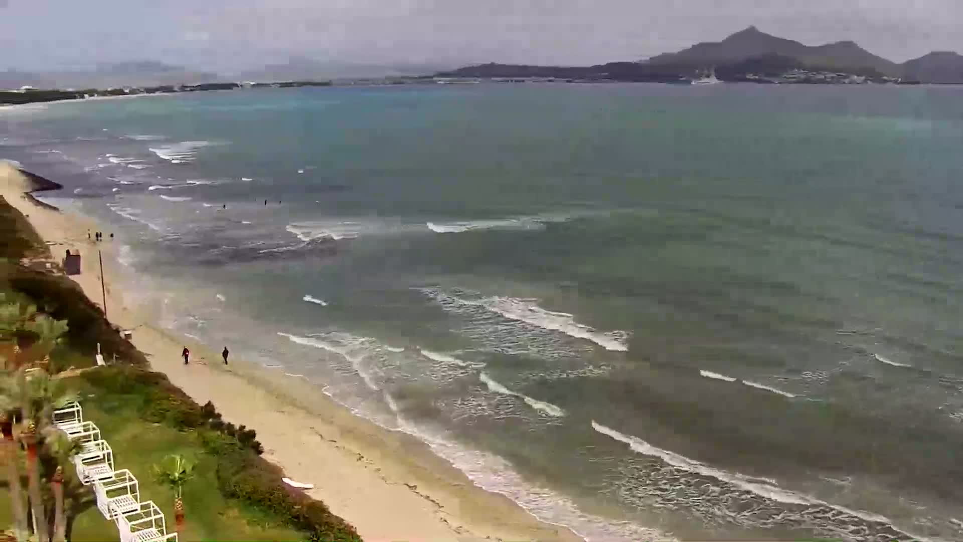 A sandy beach with a few people walking along the shore meets a calm, green-blue sea with small waves under a cloudy sky, with distant hills visible across the water.