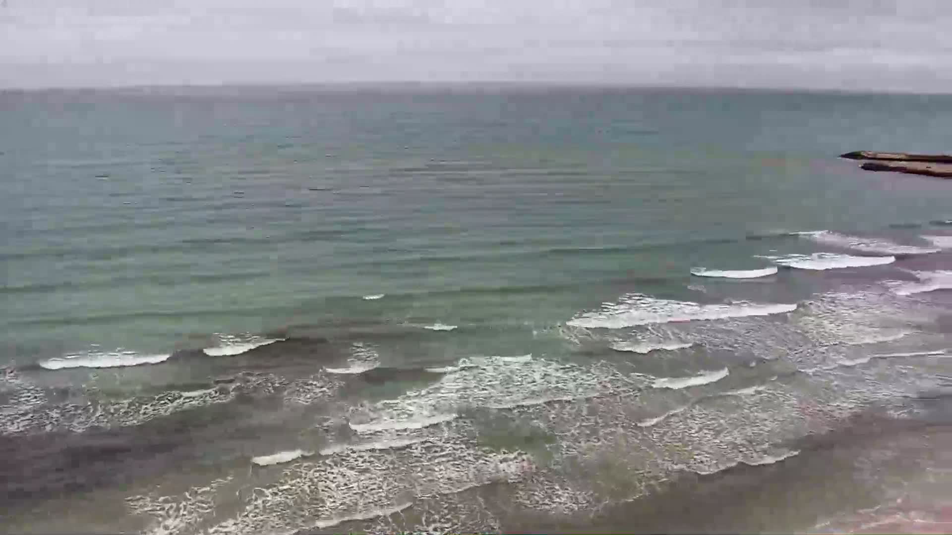 A wide shot shows a grey and green ocean with small waves breaking on a sandy shore under an overcast sky.