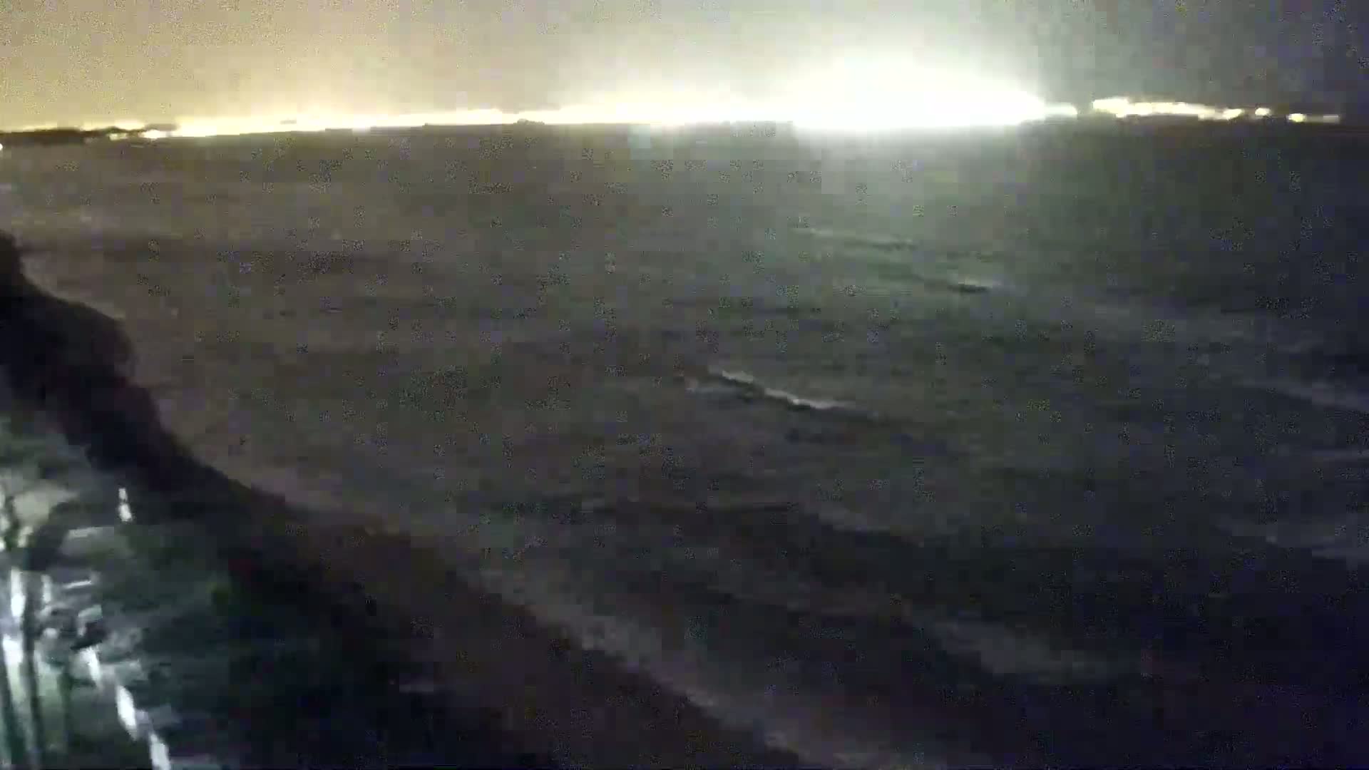 A dark, nighttime coastal scene shows a choppy sea with a distant, brightly lit shoreline.