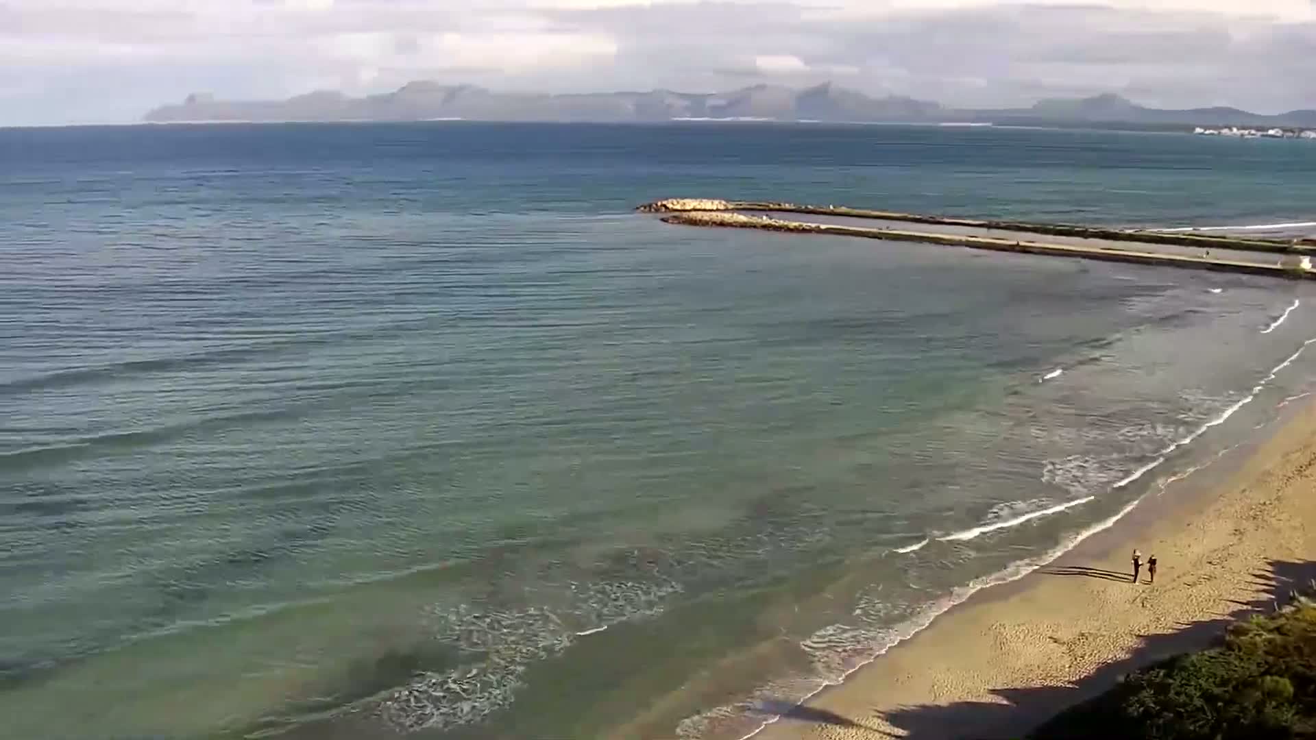 A sandy beach meets a calm, teal ocean under a partly cloudy sky, with a long jetty extending into the water and distant mountains visible on the horizon.