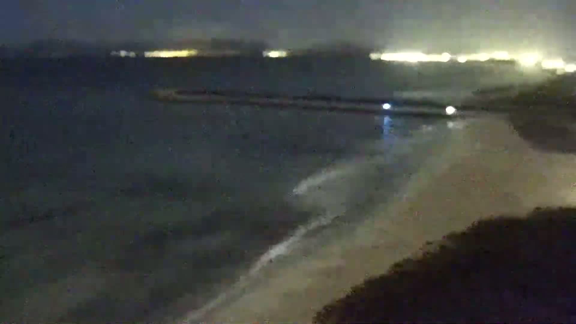 A nighttime beach scene shows a dark ocean with small waves lapping onto a sandy shore, with distant lights visible across the water.