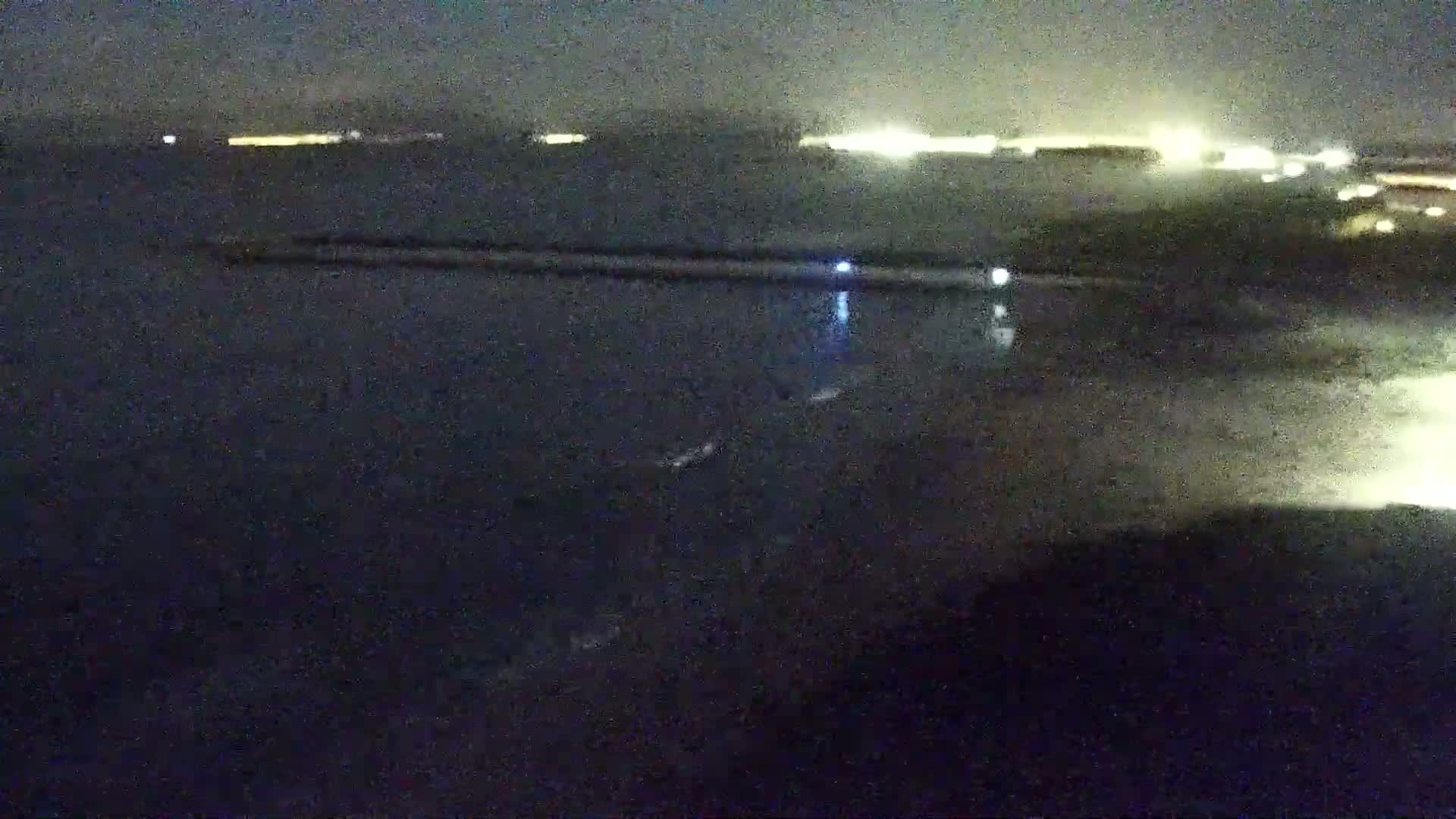 A nighttime, long shot of dark water with a long structure extending into it, faintly lit city lights in the distance.