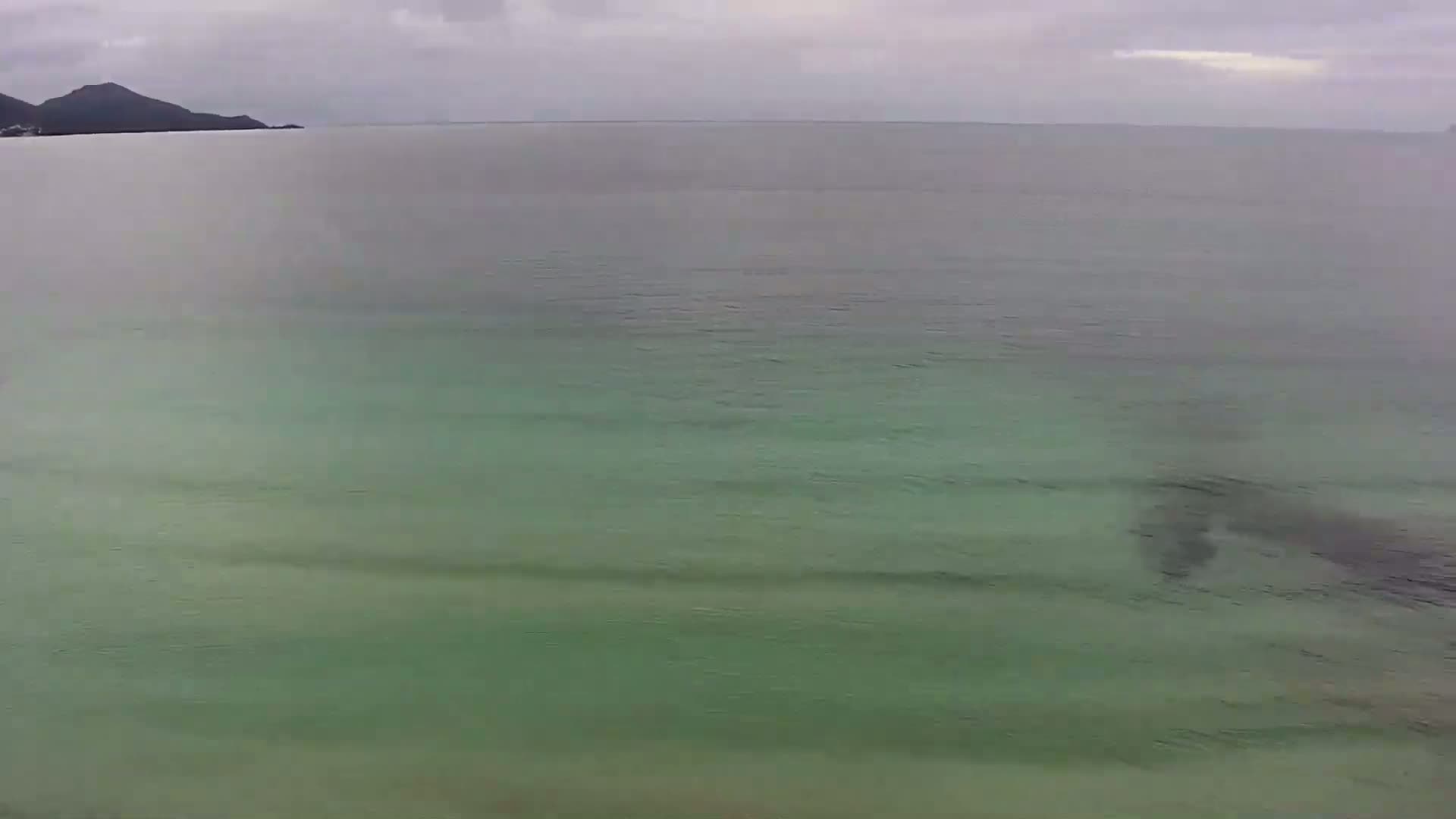 A calm, green-tinged ocean stretches to a distant, dark mountain under an overcast sky.