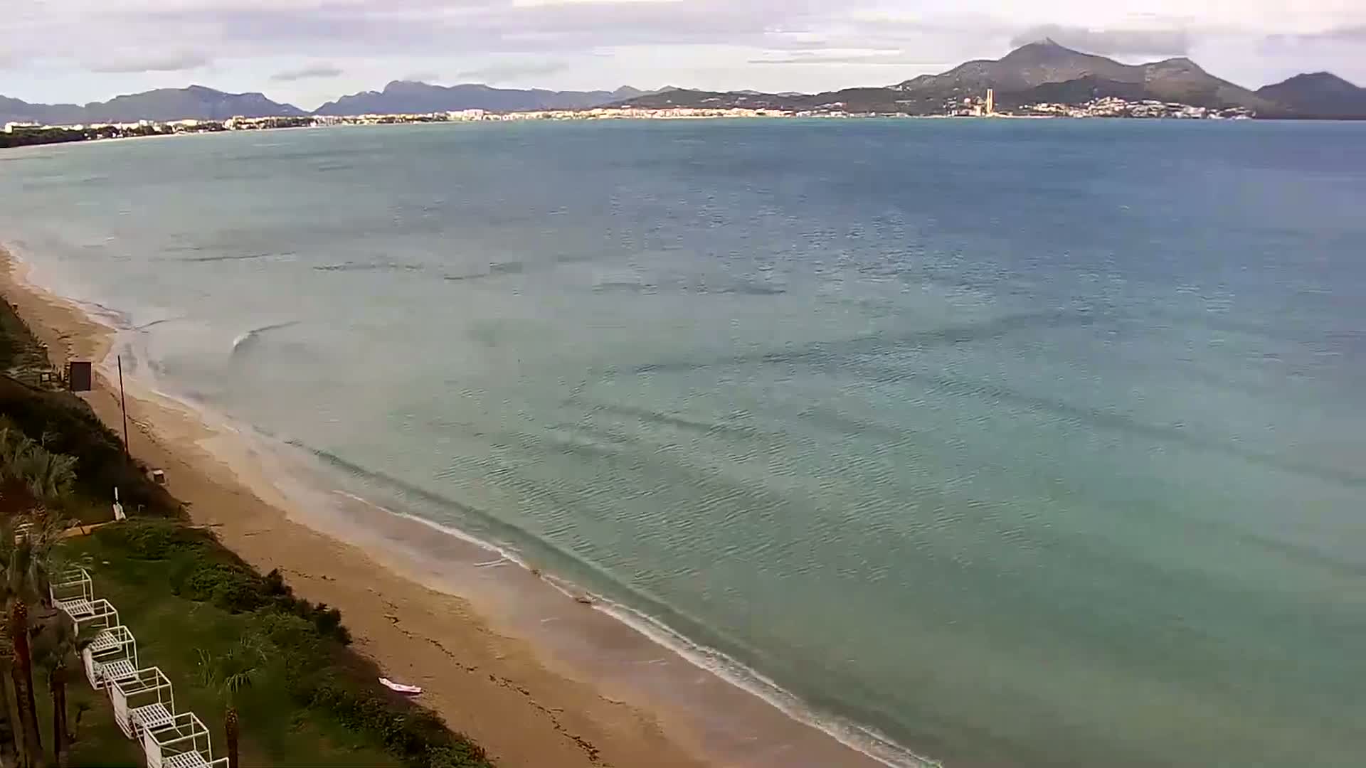 A sandy beach with gentle waves laps the shore under a cloudy sky, with mountains visible in the distance.