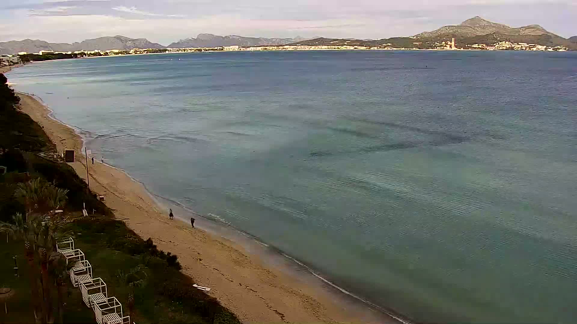 A sandy beach slopes gently into calm, teal-colored water under a partly cloudy sky, with mountains visible in the distance.