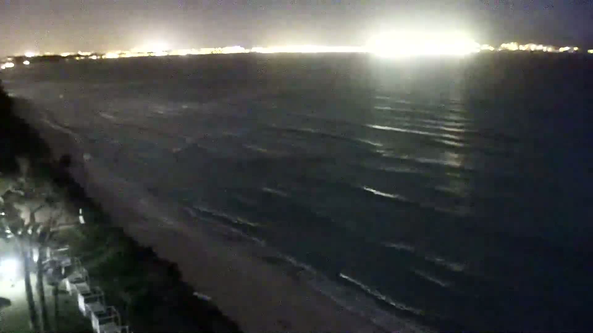 A nighttime view of a beach with small waves lapping the shore, under a dark sky illuminated by distant city lights.