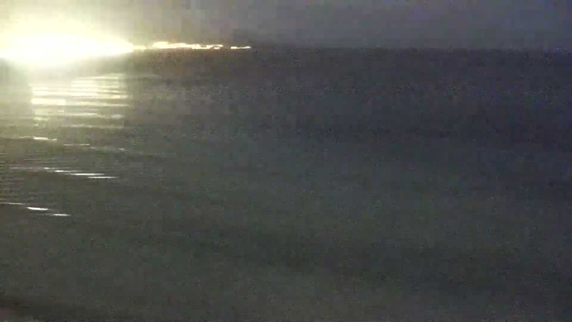 A dark, somewhat blurry image shows a body of water with gentle waves reflecting a bright light source on the horizon at night.
