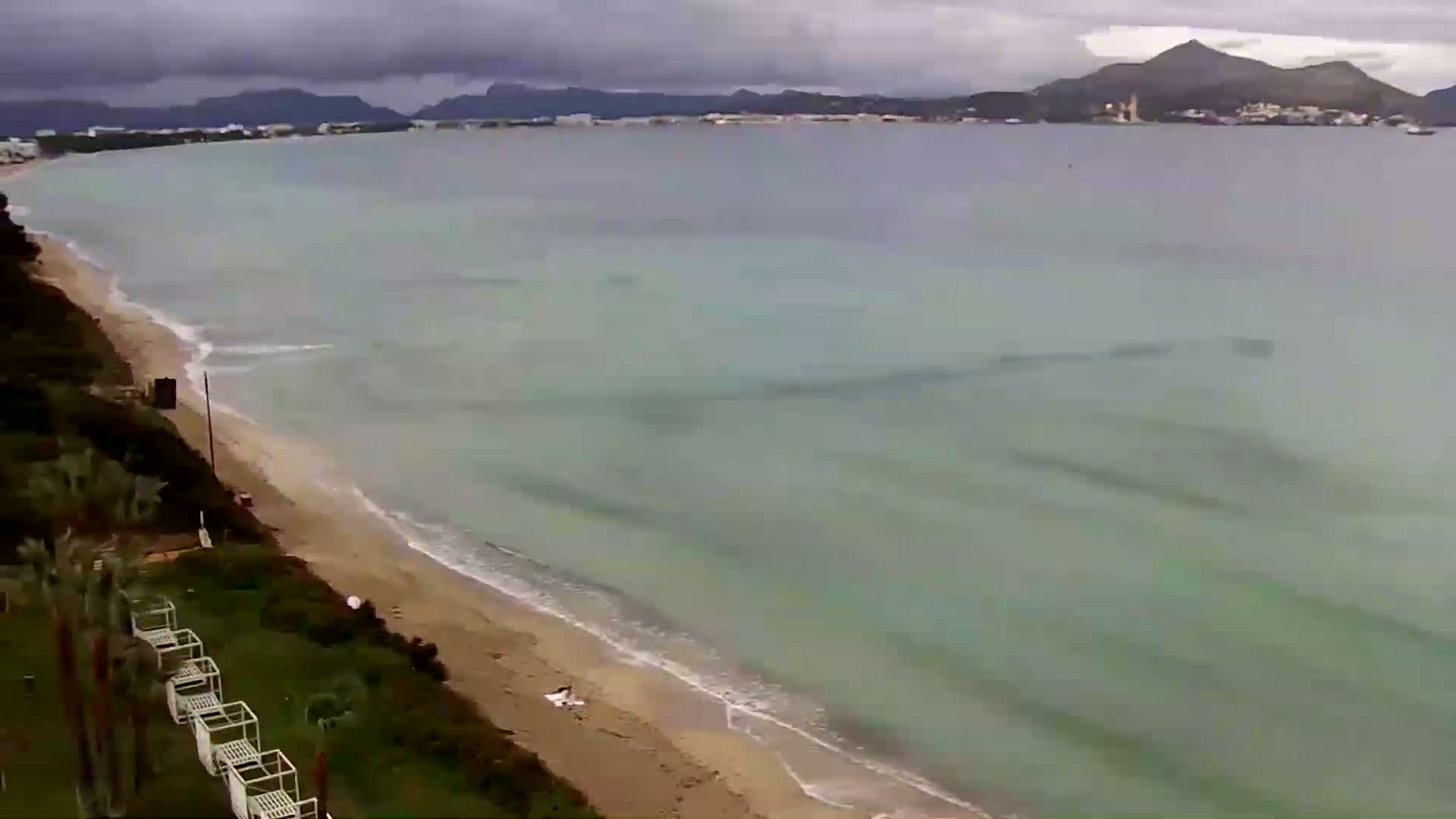 A sandy beach with calm, greenish-blue water stretches to a distant coastline under an overcast sky.
