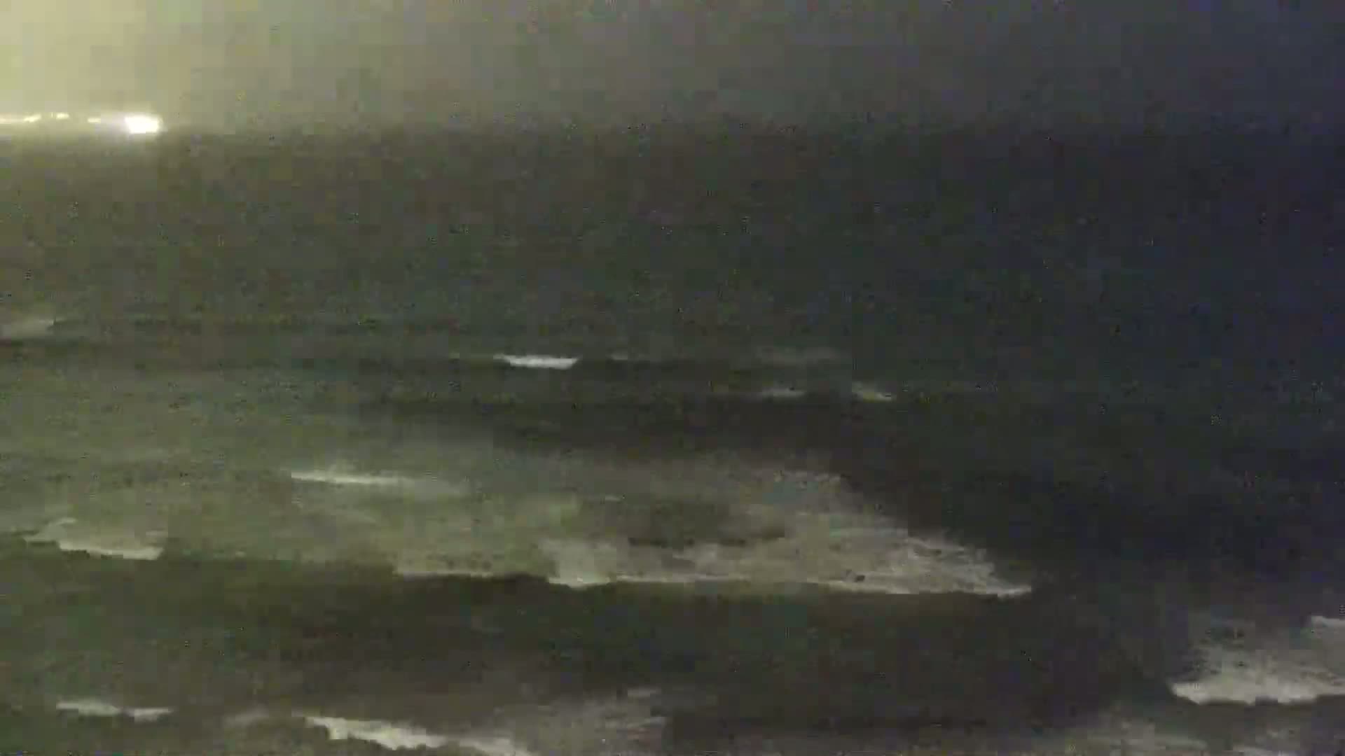 A dark, nighttime ocean scene shows waves breaking on a shore under hazy, overcast conditions.