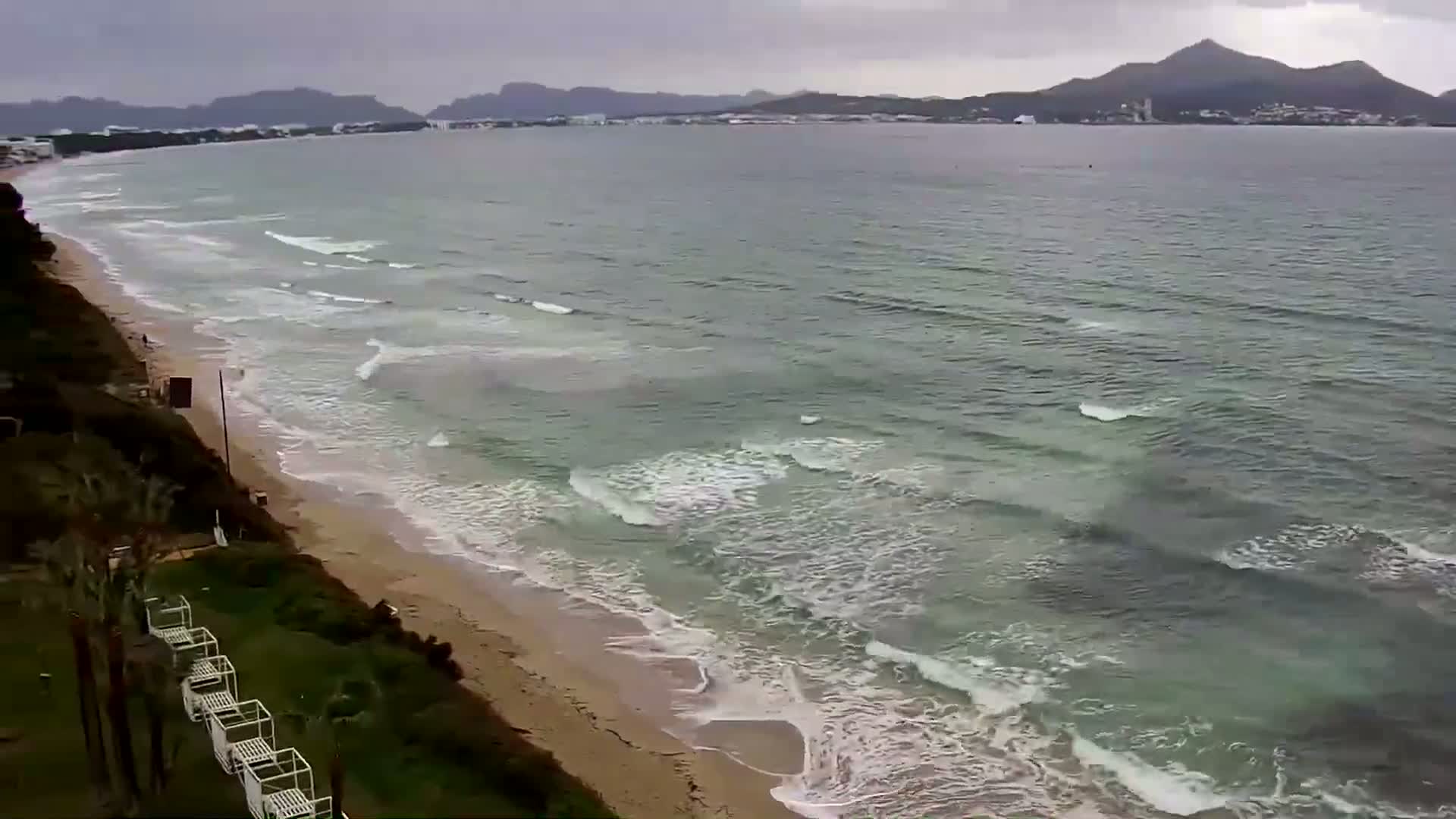A sandy beach with gentle waves rolls onto the shore under an overcast sky, with mountains visible in the distance.