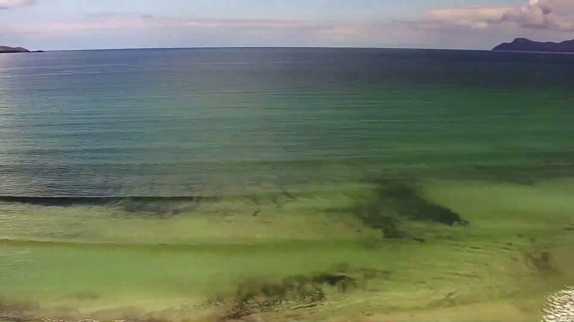 An aerial view shows a calm, shallow, green-tinged ocean with small waves lapping a sandy shore under a partly cloudy sky.