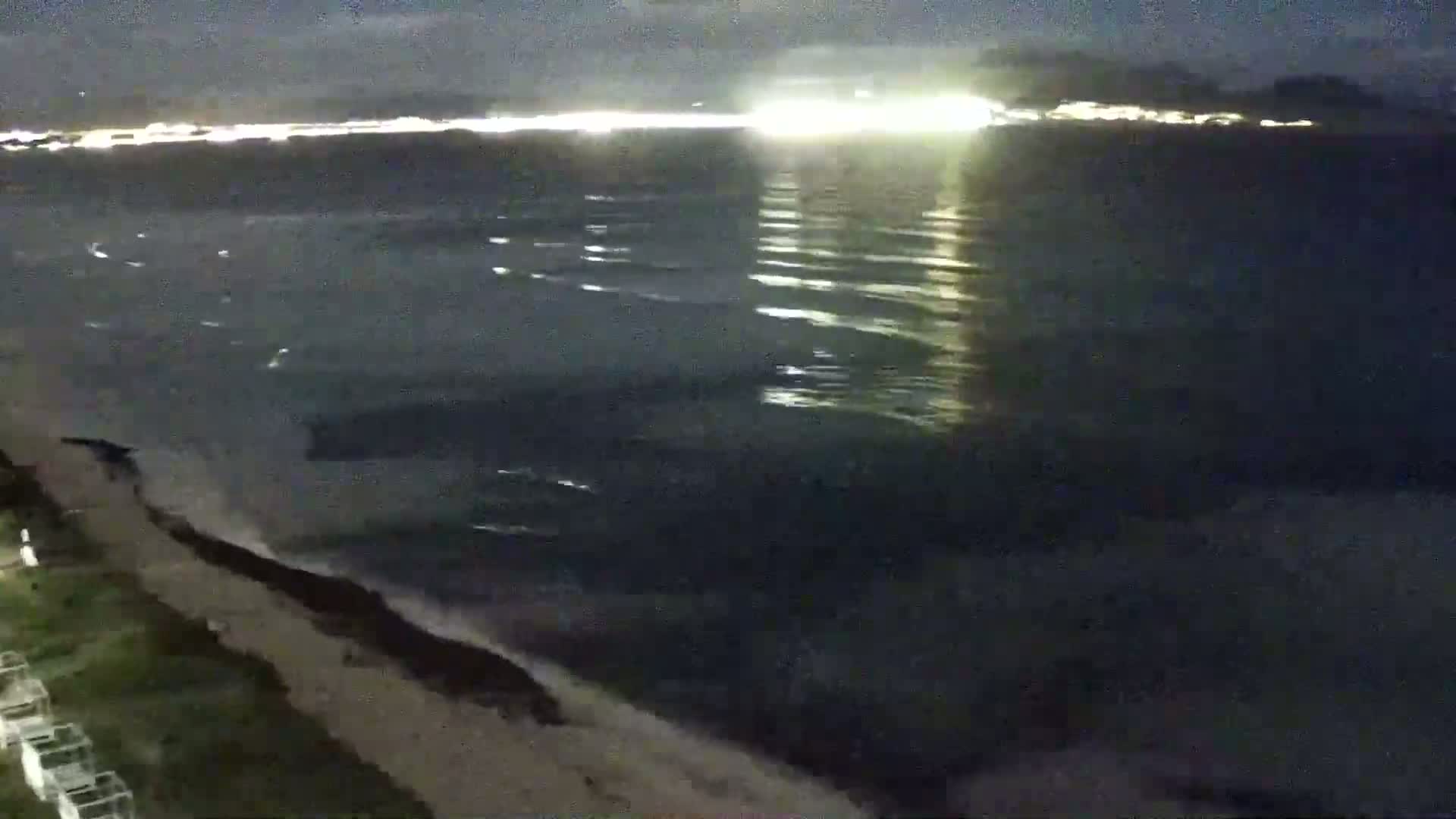 A dark, nighttime beach scene shows a choppy ocean with city lights reflecting on the water in the distance.