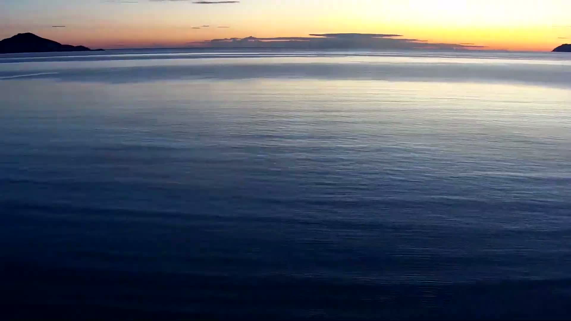 A calm, dark blue ocean stretches to the horizon under a sunset sky with silhouetted hills in the distance.