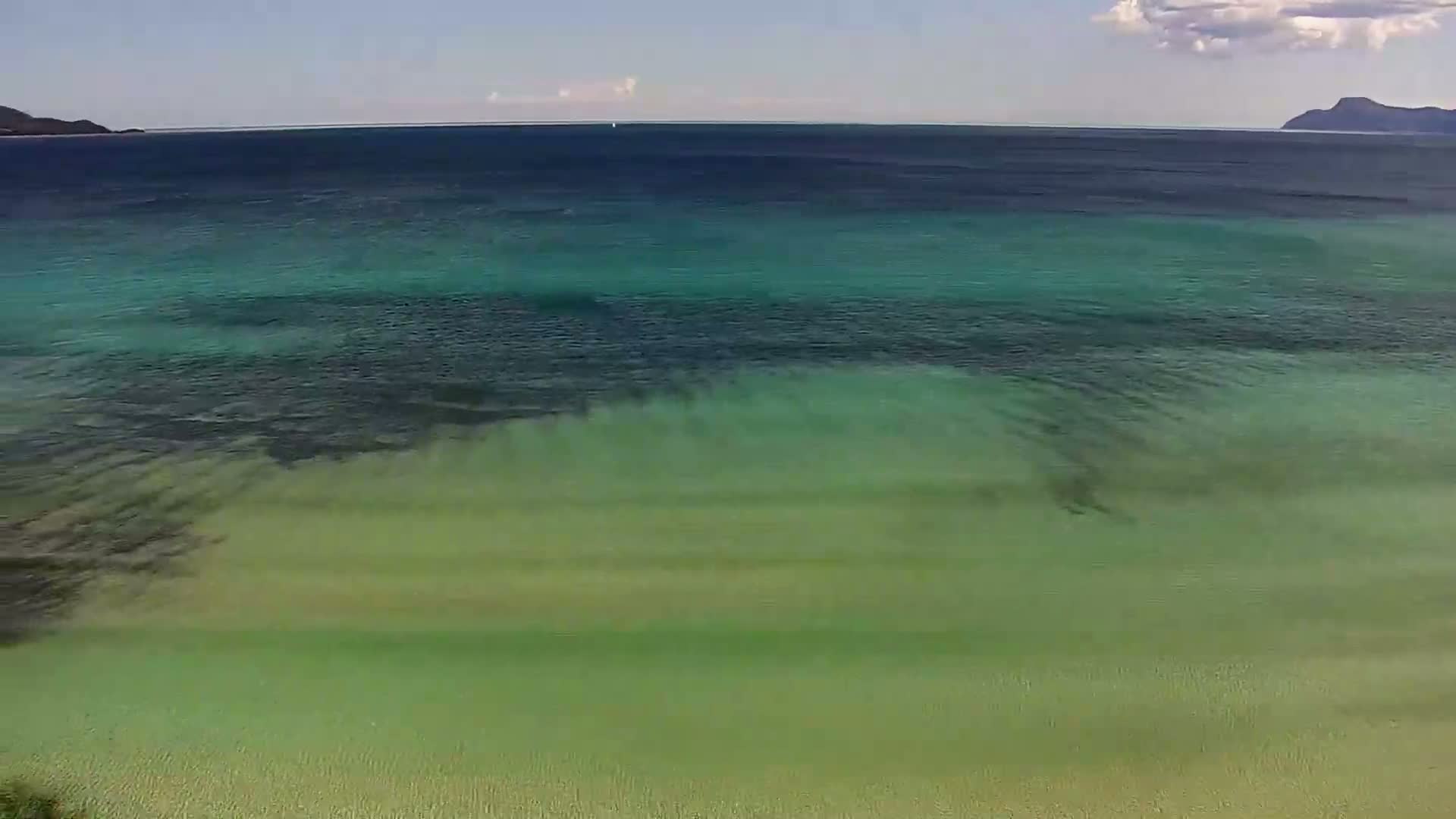 Shallow, clear, turquoise water laps a sandy shore under a mostly sunny sky with distant hills.