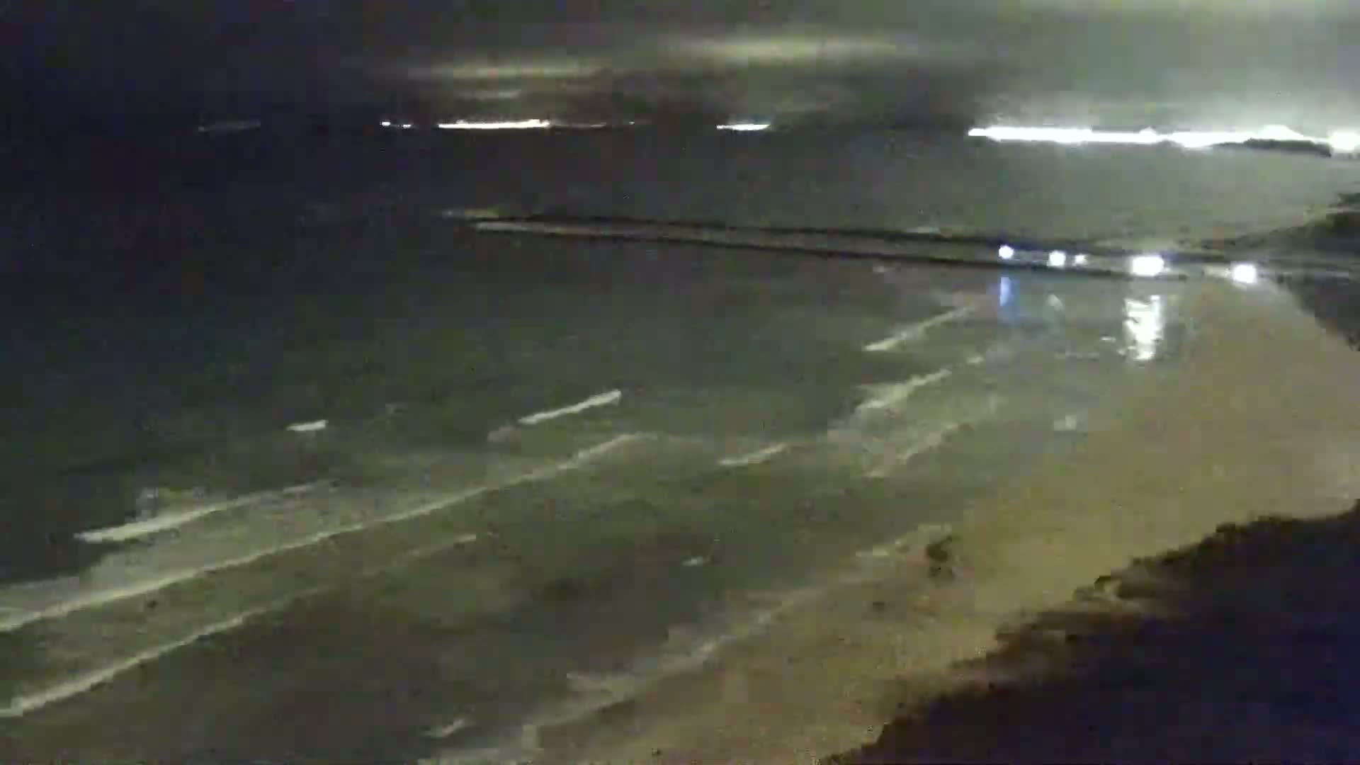 A nighttime view of a beach with gentle waves lapping the shore under a dark, cloudy sky.