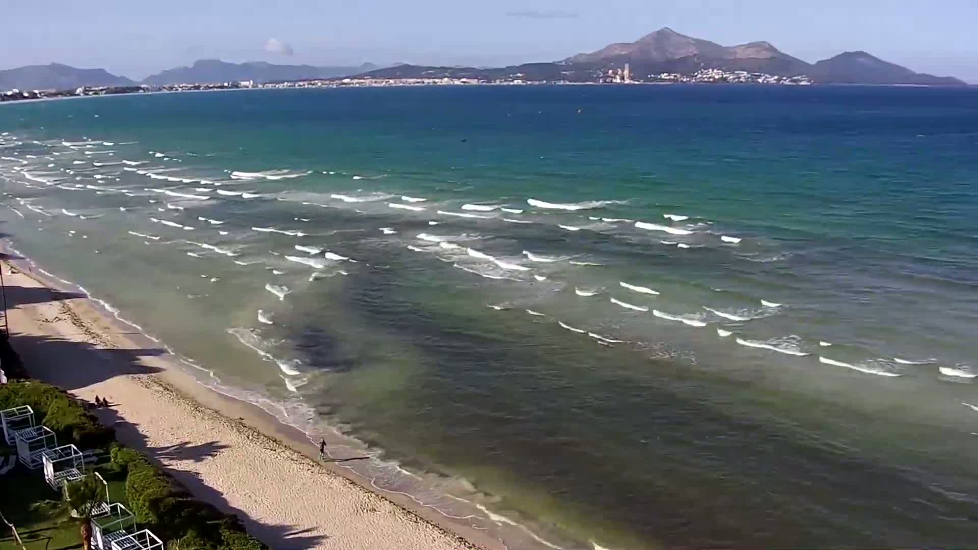 A sandy beach with small waves laps a shoreline under a clear sky with mountains visible in the distance.