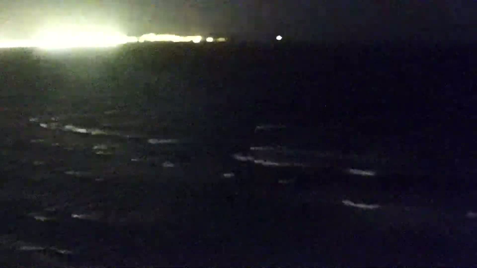 A dark, choppy sea is visible at night, with a distant line of lights on the horizon.