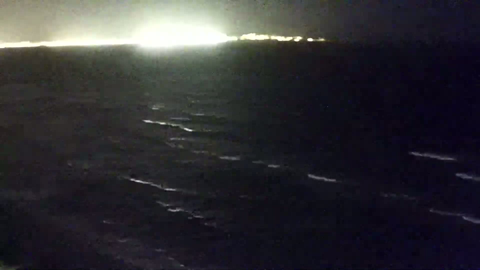 A dark, nighttime ocean with small waves is seen, with a distant, brightly lit shoreline visible on the horizon.