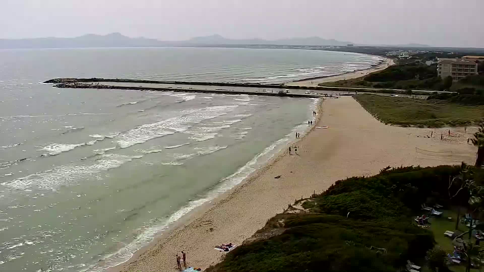 A sandy beach with a few people on it slopes down to a gently-lapping sea under an overcast sky, a low stone jetty extends into the water.