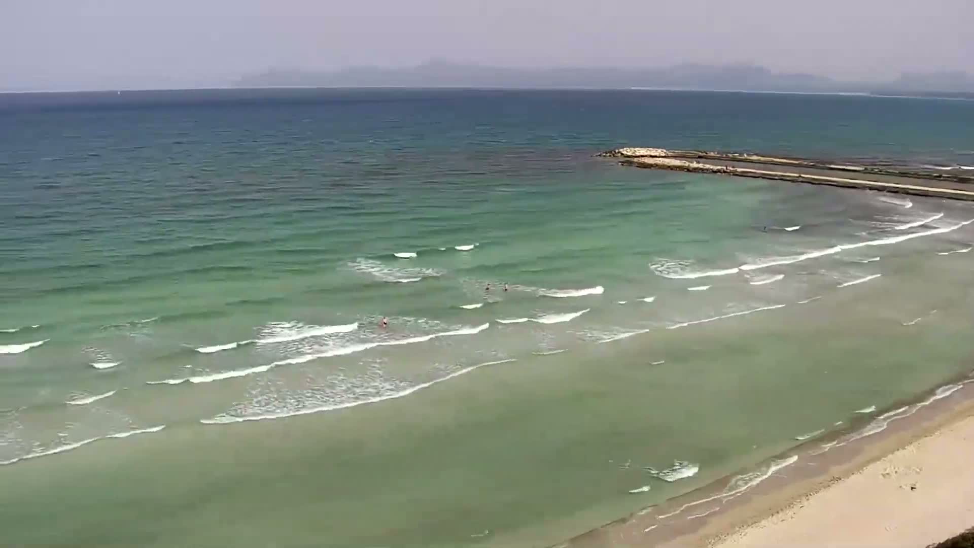 A sandy beach with light-green water and small waves laps gently against the shore under a hazy sky, a man-made structure extends into the sea.