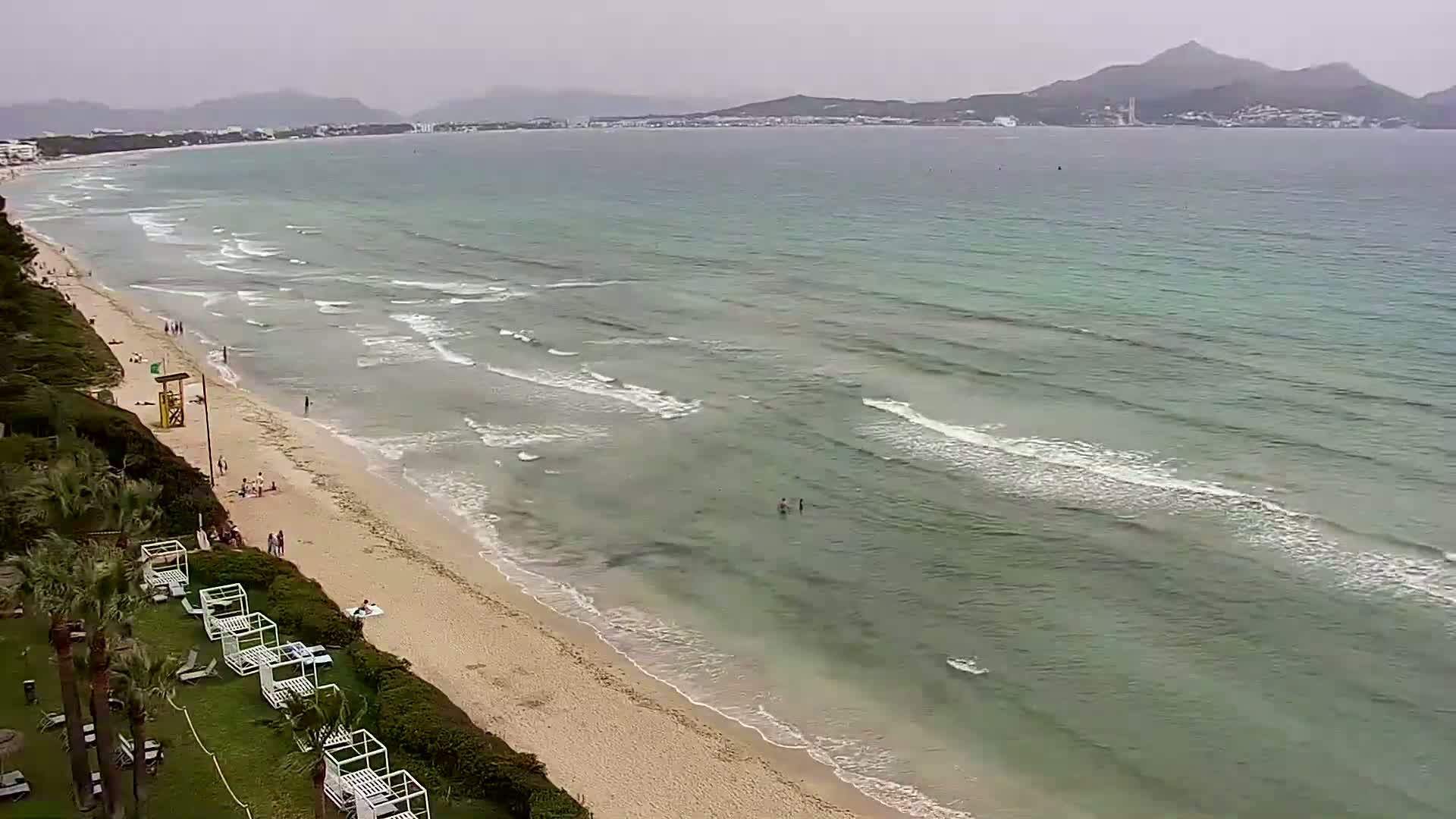 A sandy beach with a few people and white beach cabanas is seen from above, next to a calm, light-green ocean under an overcast sky, with distant hills visible across the water.