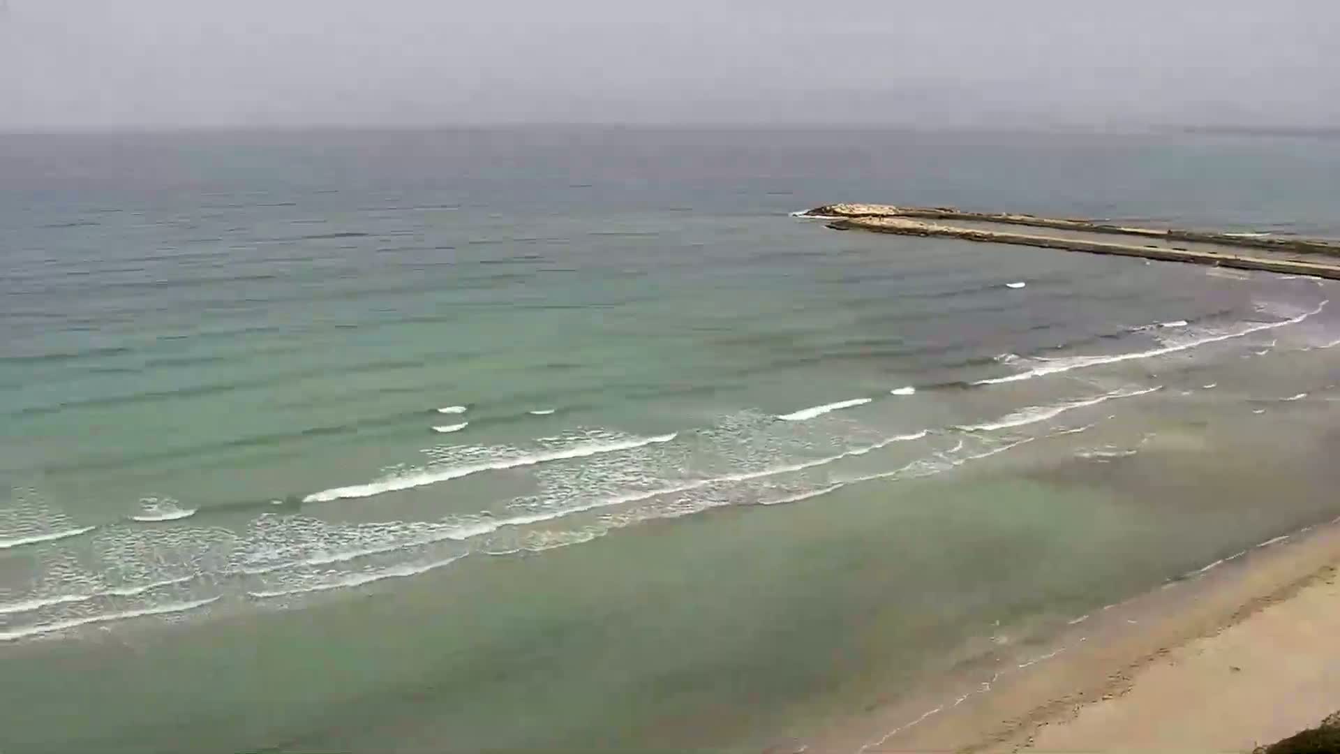 A long, sandy beach meets a calm, pale green sea with small waves under an overcast sky.