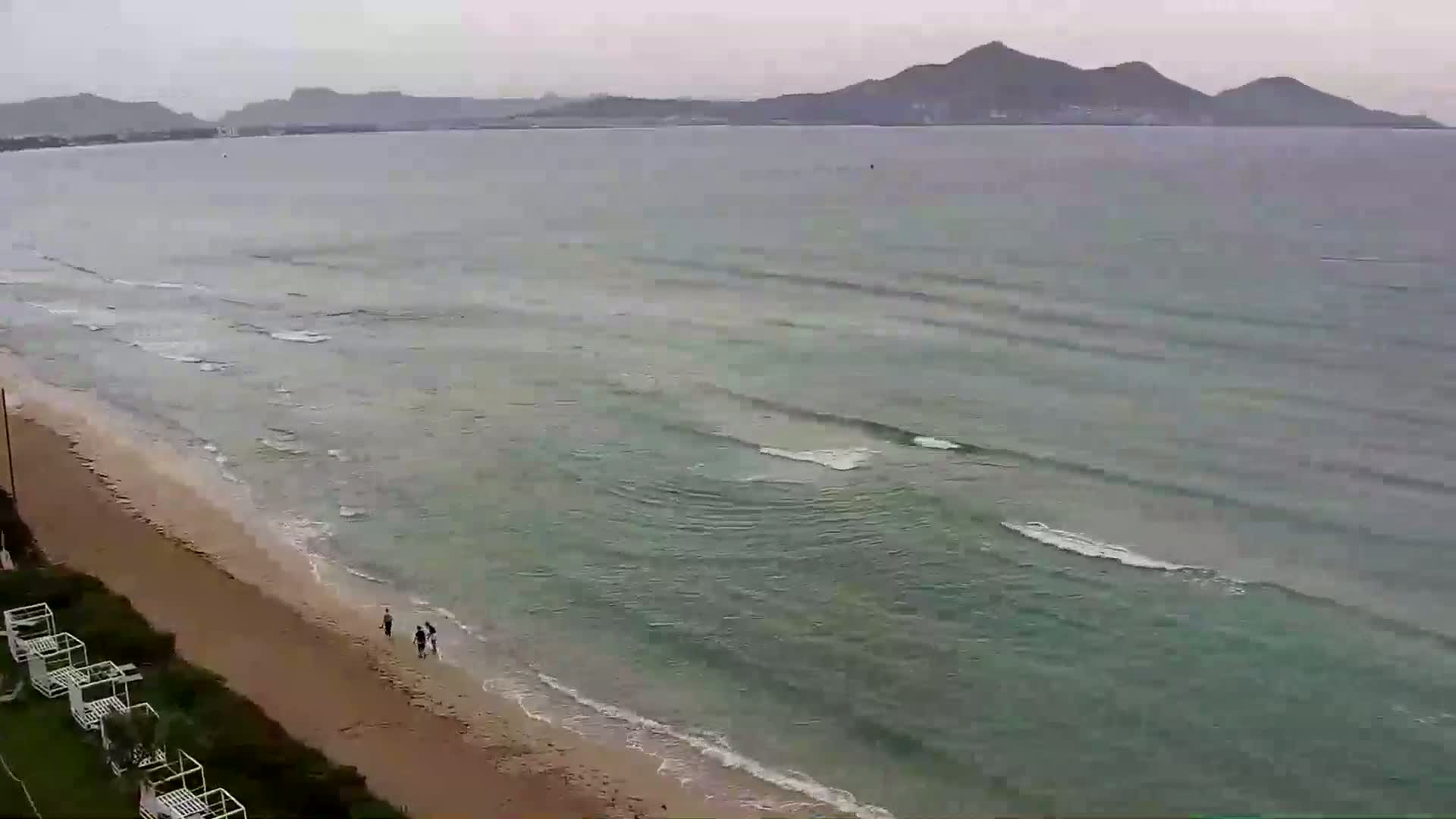 A sandy beach with a few people walking near the water's edge, gently rolling waves on a calm sea, and mountains in the distance under an overcast sky.