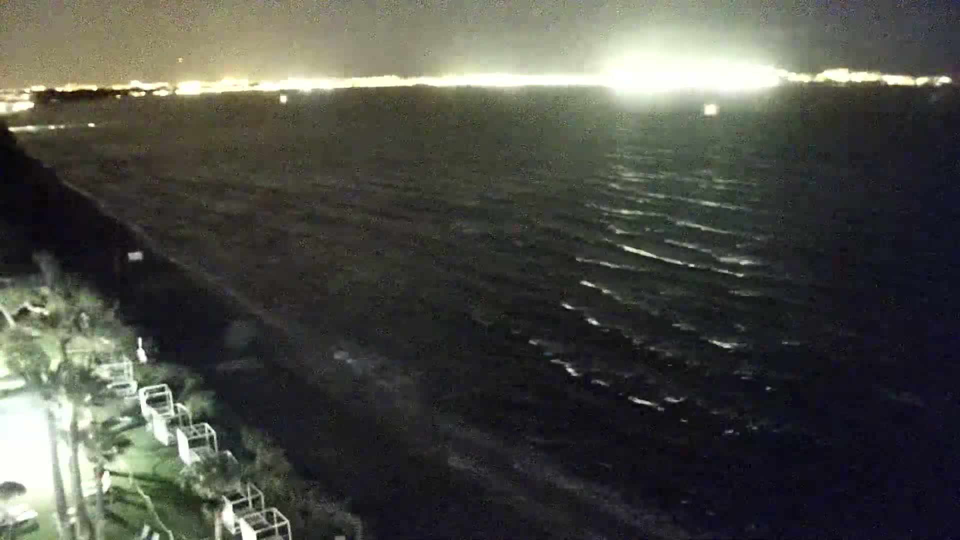 A nighttime view shows a dark, wavy ocean under a partly cloudy sky with distant city lights visible on the horizon.