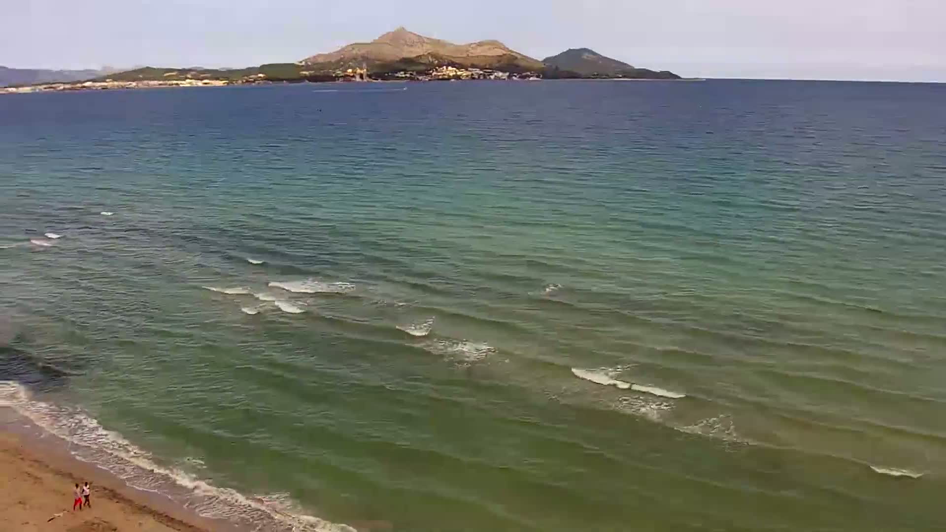 A sandy beach with two people walking along the shore, gentle waves lapping at the sand, and a calm sea extending to a distant mountain range under a clear sky.