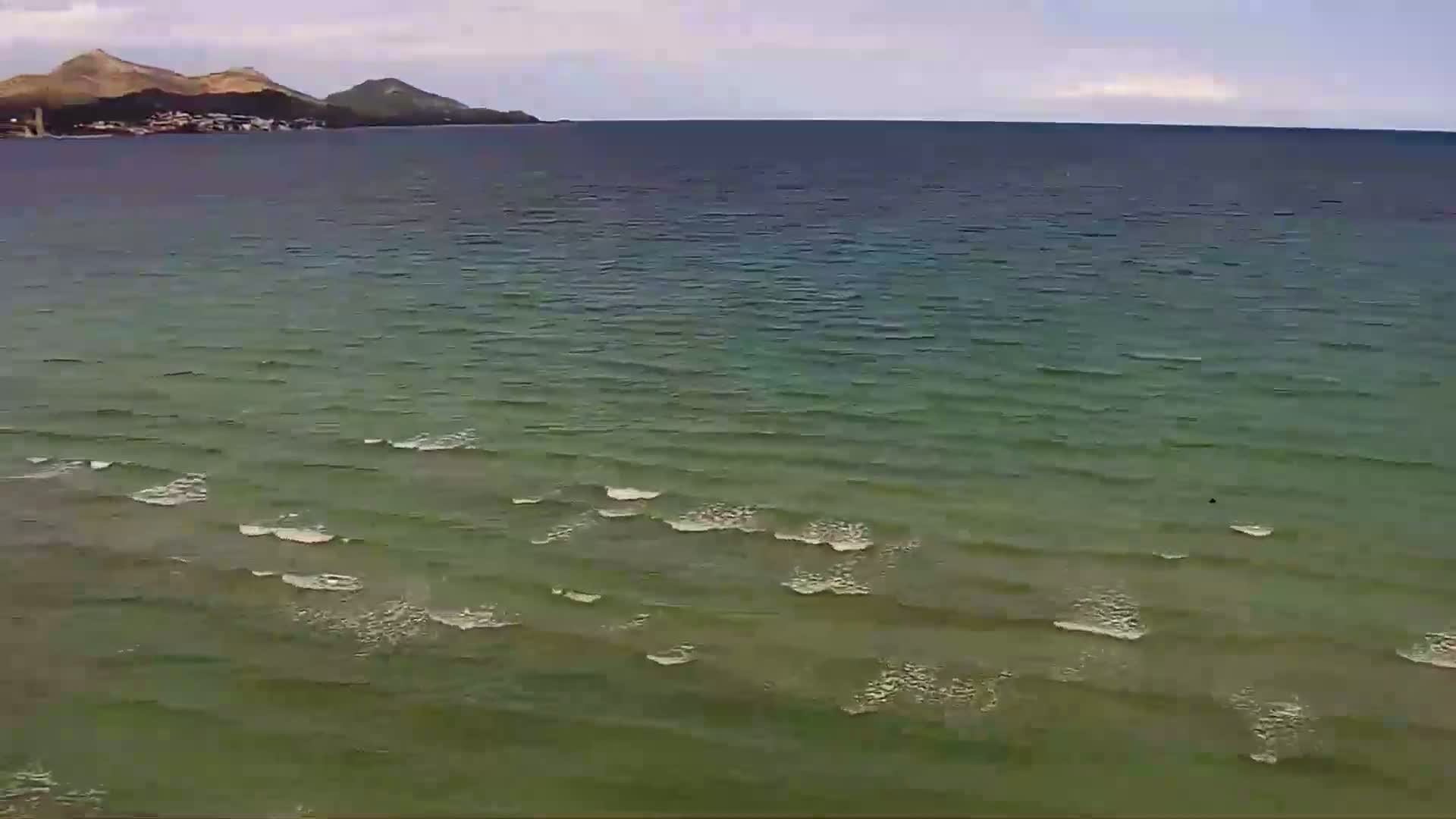 A calm sea with small waves laps gently against a shore, with distant hills visible under a cloudy sky.
