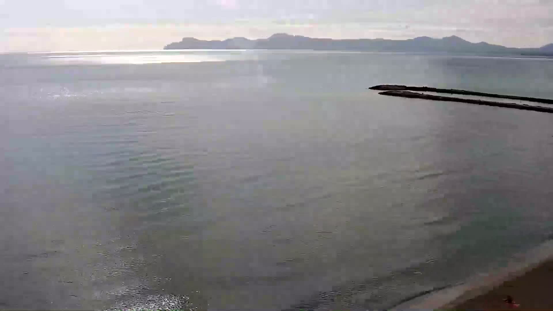 A calm, gray sea stretches to a distant mountain range under a pale sky, with a small breakwater visible in the foreground.