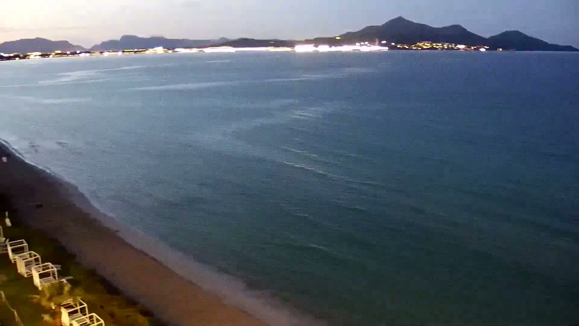 A sandy beach slopes gently into a calm, dark-green sea under a twilight sky, with a distant city's lights visible across the water.