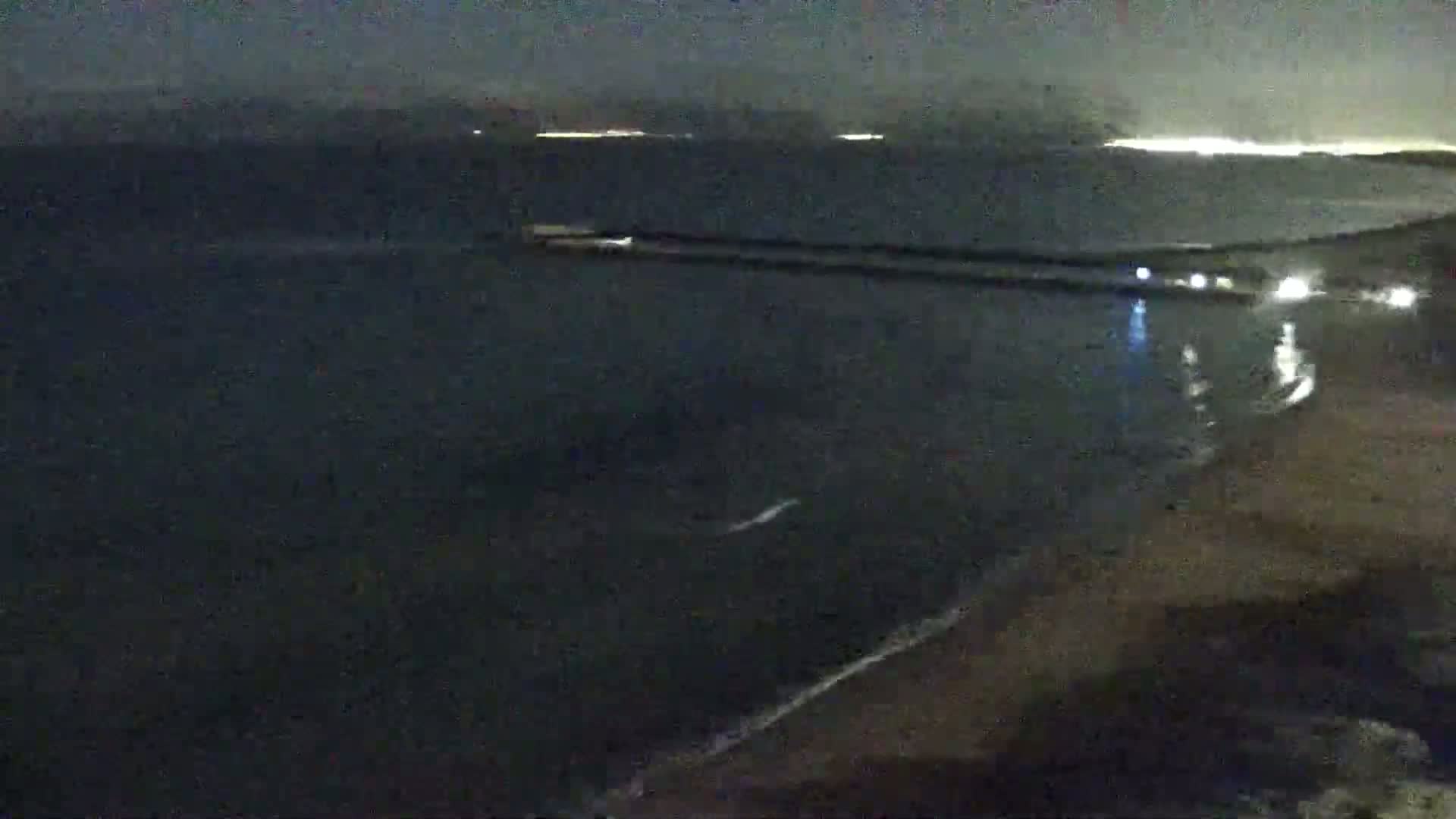 A nighttime coastal scene shows dark water with small waves lapping a sandy shore under a dark sky with distant city lights.