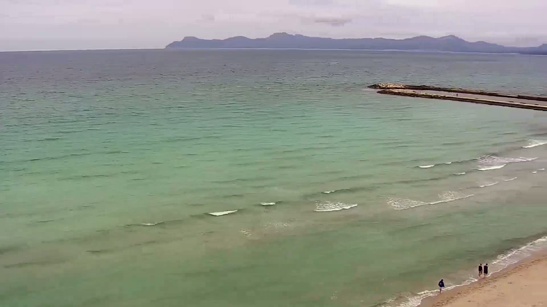 An aerial view shows a sandy beach with a few people walking near the water's edge, gentle waves lapping at the shore, and a calm, light-green sea extending to a distant mountain range under a cloudy sky.