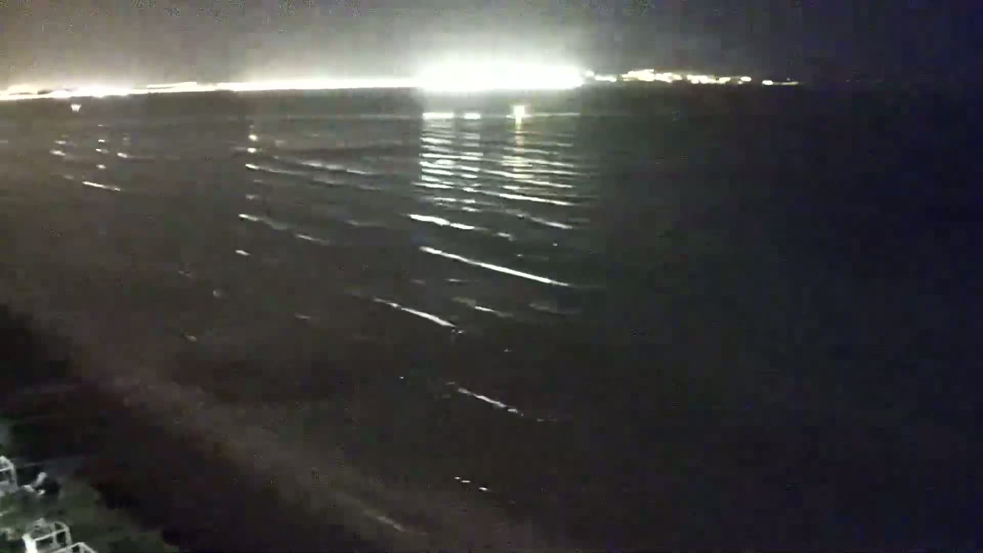 A dark, nighttime view of a gently rippling body of water with a distant, brightly lit shoreline.
