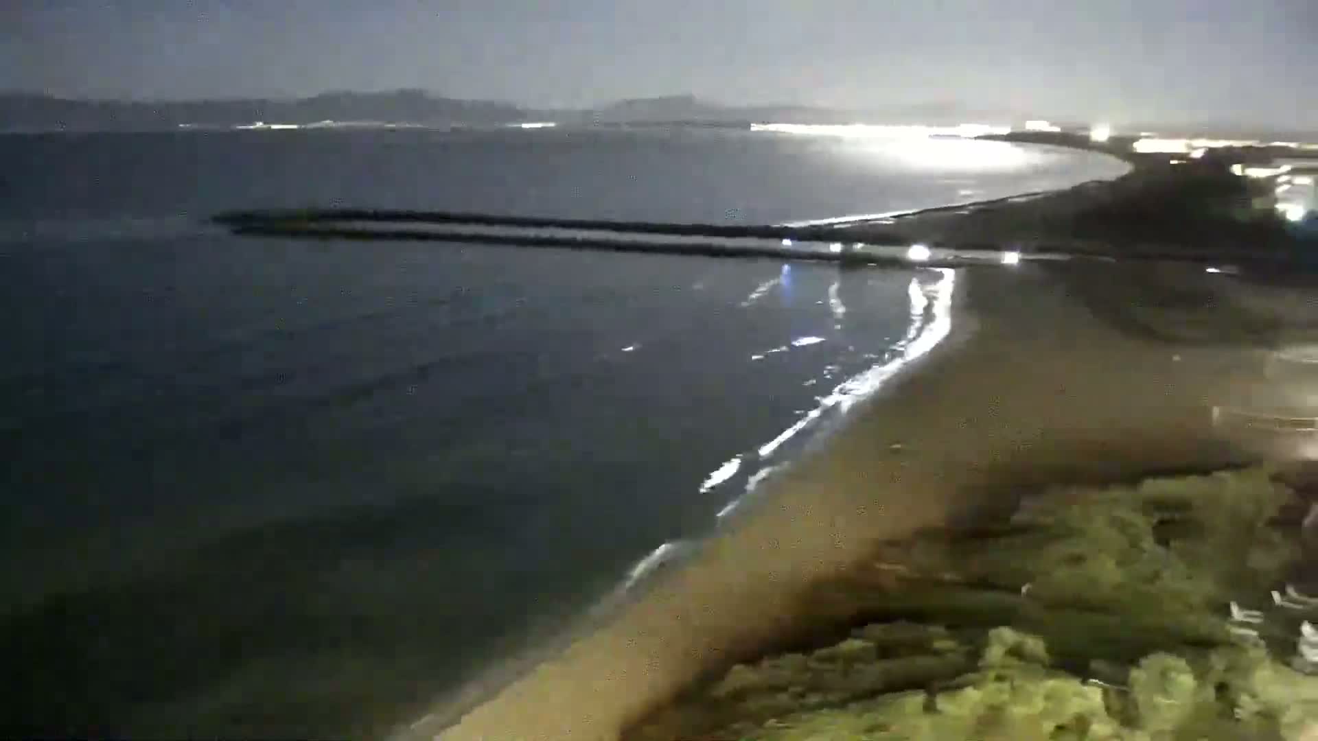 A nighttime view shows gently lapping waves on a sandy beach, with a dark body of water extending to a distant, faintly lit coastline.