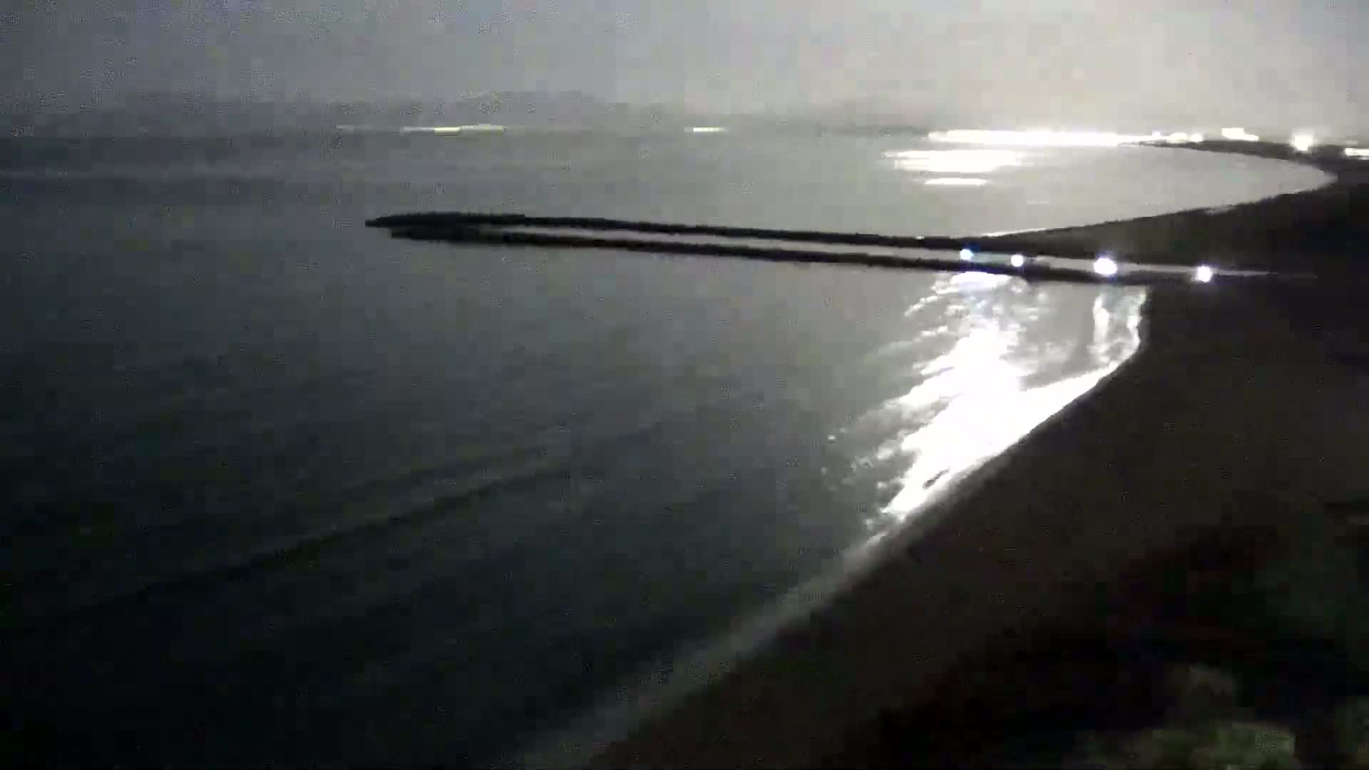 A dark beach at night with gentle waves lapping the shore, a long breakwater extending into the calm water, and faint lights visible in the distance.