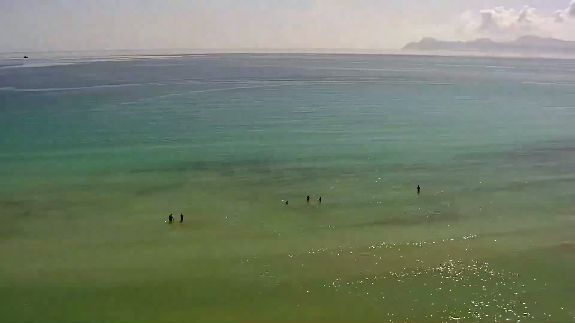 A calm, clear, teal-green ocean with several people wading in shallow water near the shore, a distant coastline visible on the horizon under a mostly clear sky.