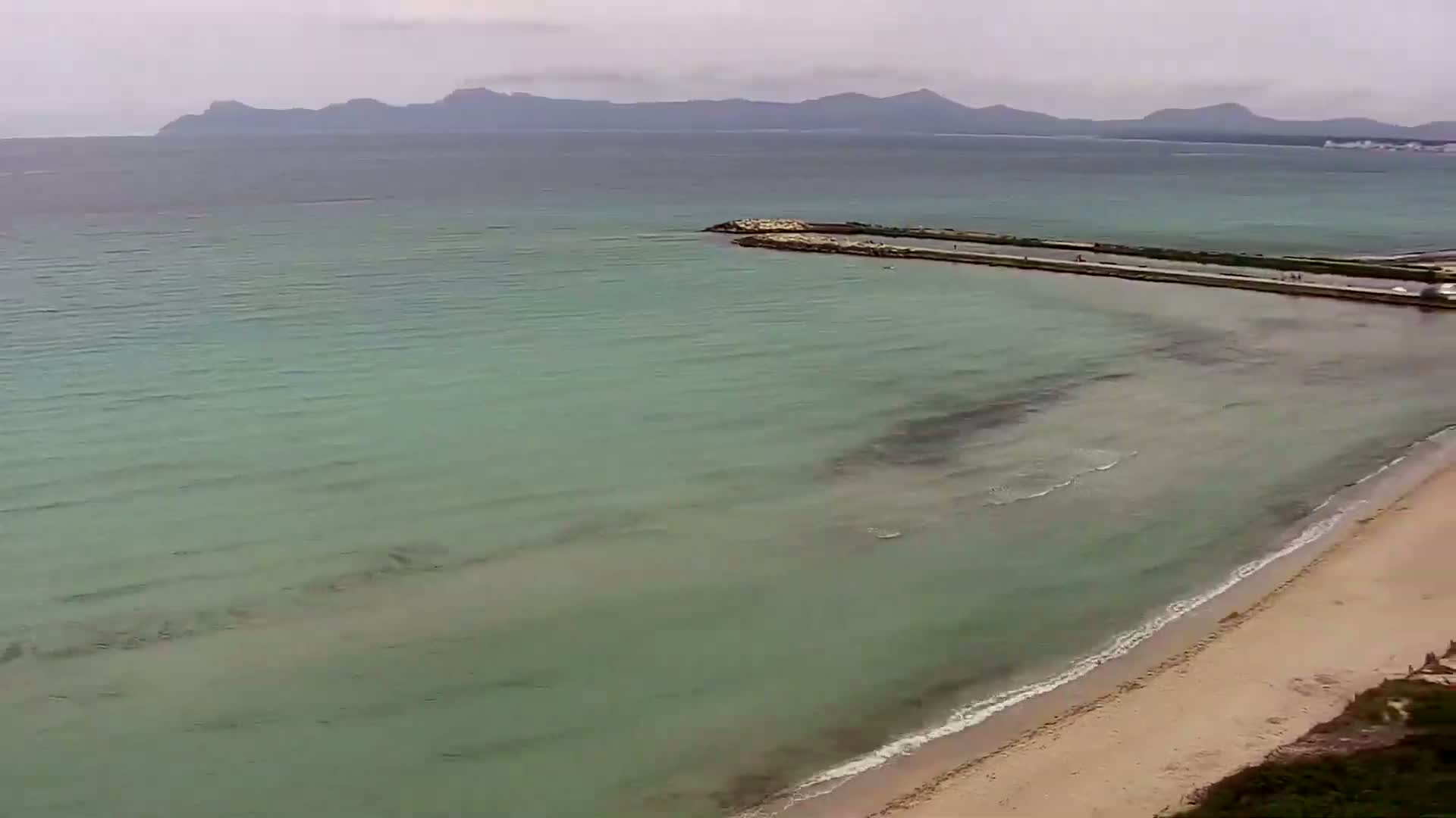 A light-green sea laps a sandy beach under an overcast sky, with mountains visible in the distance.