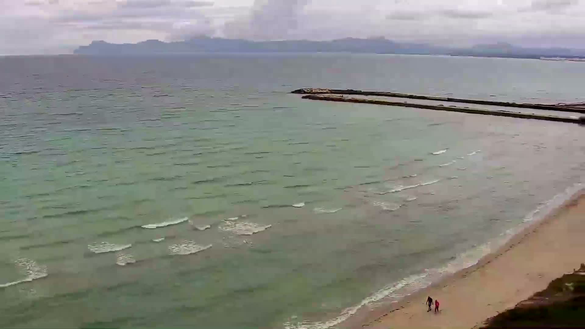 An overcast day shows a calm sea lapping a sandy beach with two people walking near the water's edge, and a distant mountain range visible on the horizon.