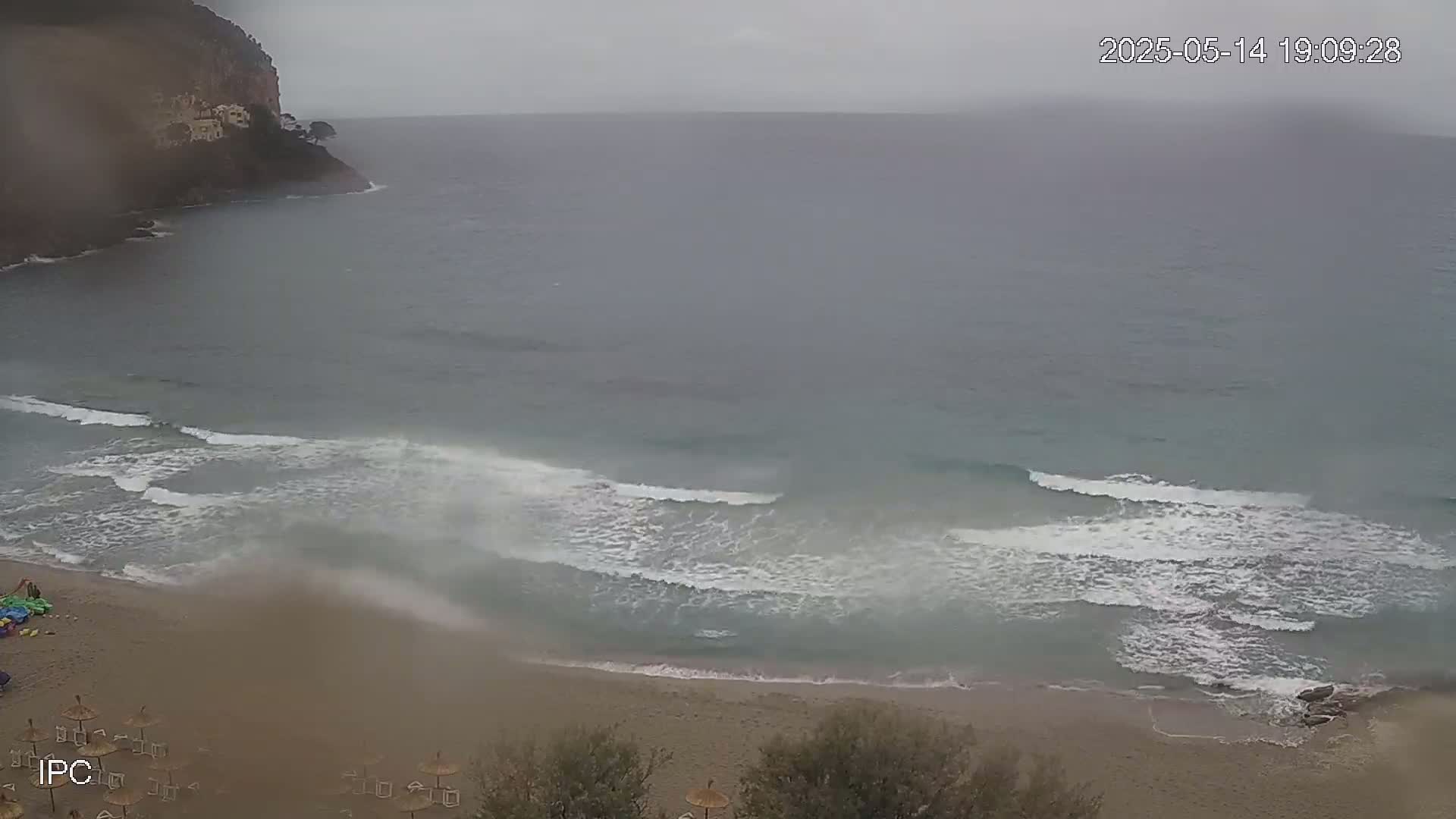 A sandy beach with small waves and a rocky cliff in the background on an overcast day.
